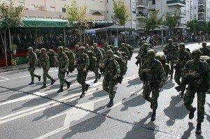 Soldados em camuflado marcham em coluna numa rua urbana com edifícios residenciais e multidão de espectadores ao fundo.