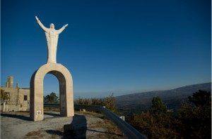 Monumento em pedra clara com arco e figura com braços levantados no topo, paisagem montanhosa ao fundo.