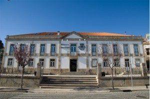 Fachada de edifício público de dois pisos com azulejos azuis e brancos, telhado de telha cerâmica, escadaria central e árvores laterais.
