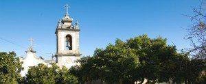 Igreja branca com campanário de cúpula, cruz no topo e sino, rodeada de vegetação.