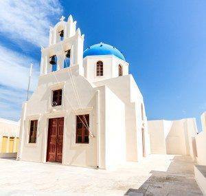 Igreja branca com cúpula azul, campainha de sino e portas vermelhas, sob céu azul claro.