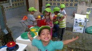 Crianças em coletes amarelo-fluorescente e bonés verdes participam em atividade educativa com materiais de construção e segurança num pátio.