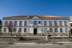 Edifício institucional de dois pisos com fachada azul e branca, escadas de acesso frontal e brasão no centro
