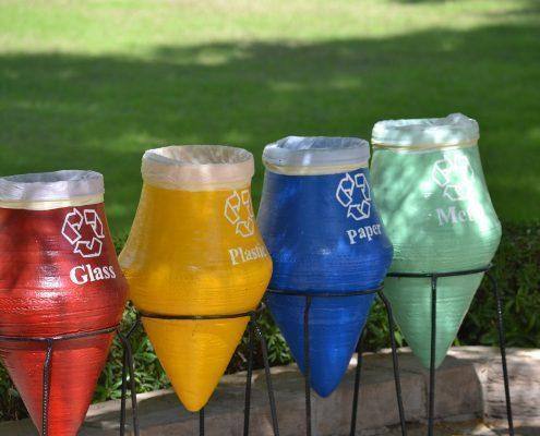 Quatro cones de reciclagem coloridos com rótulos para vidro, plástico, papel e metal, sobre estacas no jardim — ampliar imagem