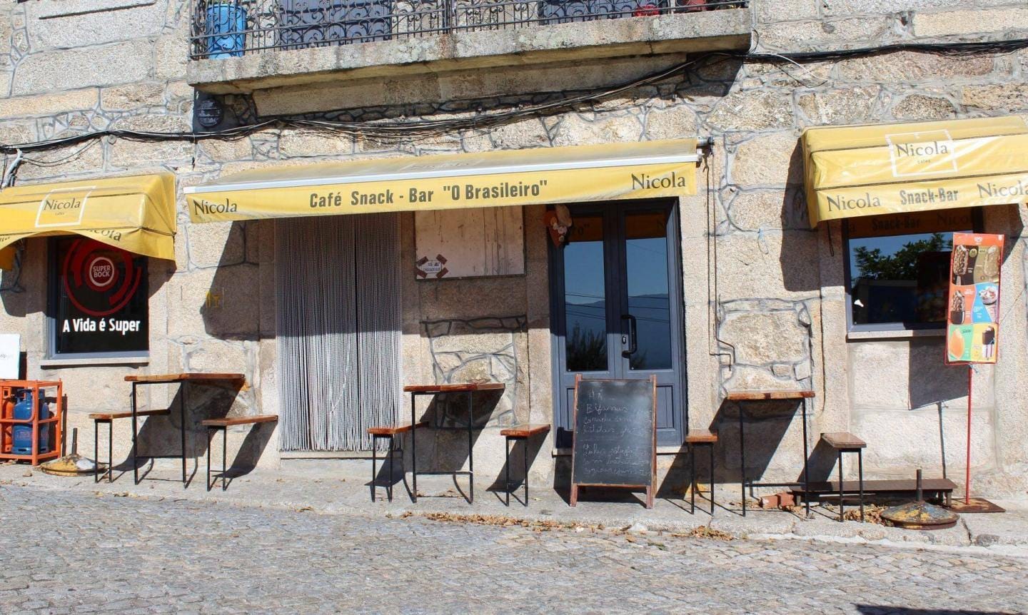 Fachada de café com toldo amarelo, cadeiras pretas e porta azul numa rua em calçada portuguesa