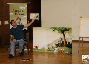 Homem sentado mostra ilustração de livro infantil junto a painel e cenário com árvore e animais