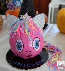Abóbora rosa pintada com rosto, antenas prateadas e crina colorida, sobre base preta com texto Halloween.