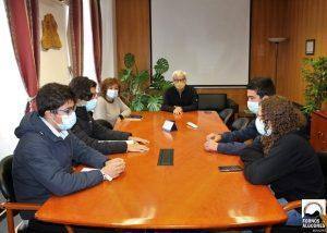 Sete pessoas sentadas à volta de uma mesa de madeira num sala de reuniões com plantas e ecrã de projeção ao fundo.
