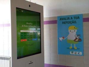 Quiosque com ecrã tátil verde para avaliar refeições e painel informativo azul com ilustração.