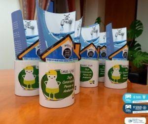 Copos brancos com mascote amarela e embalagens de cartão com torneira e água, sobre superfície laranja, com plantas de fundo.