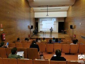 Auditório com audiência dispersa e dois apresentadores no palco junto a projetor