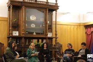 Cinco mulheres sentadas numa sala com mobiliário de madeira e paredes amarelas durante uma conferência.