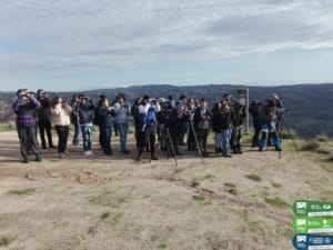 Grupo de pessoas com equipamento de observação em miradouro com paisagem montanhosa ao fundo.