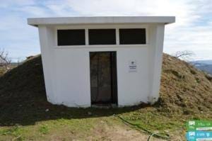 Pequeno edifício branco com três janelas e porta, situado em encosta com vegetação.