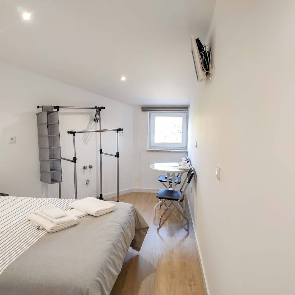 Quarto luminoso com cama de casal, cabideiro suspenso, tábua de engomar e secretária junto à janela.