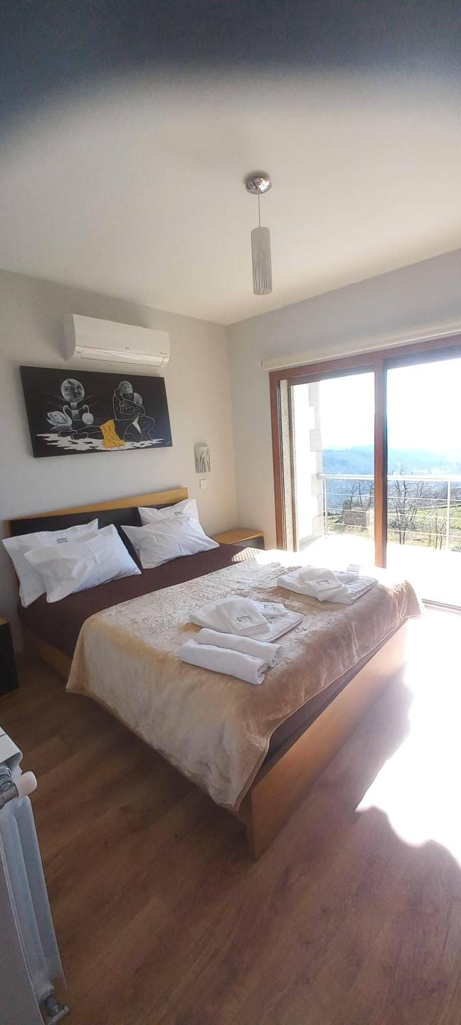 Quarto com cama de casal, mesinha lateral, quadro decorativo preto na parede, ar condicionado e porta de vidro com vista para paisagem montanhosa.