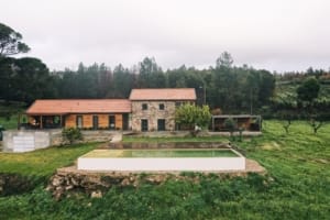 Casa moderna com fachada em pedra e laranja, piscina branca, envolvida por vinhas e floresta