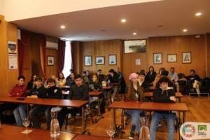 Grupo de jovens sentados em mesas de madeira numa sala de conferência, observando apresentação.