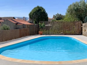Piscina azul redonda com deck em cimento, cercada por vedação em palha e árvores, edifícios rurais ao fundo.