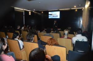 Sala de apresentação com audiência sentada e orador de pé perante ecrã de projeção