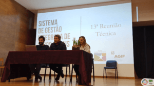 Três pessoas sentadas a uma mesa em painel numa sala de conferência com apresentação em ecrã