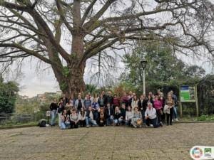 Grupo de cerca de 40 pessoas posicionado em frente a árvore antiga junto a construção de tijolo rosa