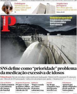 Barragem com comportas abertas e água em queda, paisagem montanhosa ao fundo sob céu nublado.