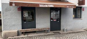 Fachada de estabelecimento com toldo castanho, nome Tasca da Terra em branco, porta preta dupla e janelas pequenas, parede branca, piso em calçada portuguesa.
