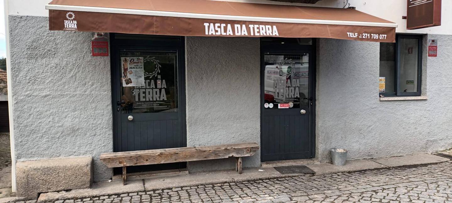 Fachada de estabelecimento com toldo castanho, nome Tasca da Terra em branco, porta preta dupla e janelas pequenas, parede branca, piso em calçada portuguesa.
