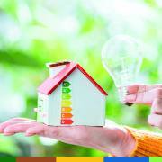 Mão segurando casa miniatura com rótulo de eficiência energética e lâmpada acesa contra fundo verde desfocado