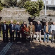 Cerimónia de homenagem com militares e coroa de flores Nove homens em pé seguram coroa de flores vermelhas perante veículo militar e muro de pedra.