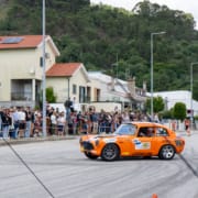 Automóvel laranja clássico em movimento numa rua durante evento com espectadores.