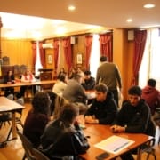 Reunião pública Dia da Europa Grupo de pessoas em reunião numa sala com mesas de madeira, cortinas vermelhas e bandeiras.