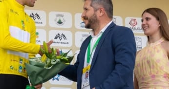 Ciclista de amarelo recebe buquê de flores de homem em traje durante cerimónia de premiação