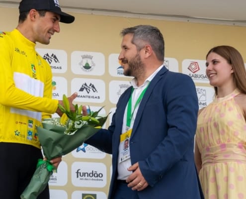 Ciclista de amarelo recebe buquê de flores de homem em traje durante cerimónia de premiação