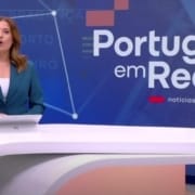 Apresentadora de informação em estúdio RTP1 com cenário Portugal em Rede ao fundo