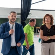Homem de blazer azul com microfone e mulher de cabelo ondulado durante evento em pavilhão.