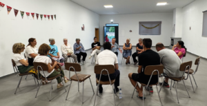 Grupo de pessoas sentadas em círculo durante sessão de informação em sala comunitária.